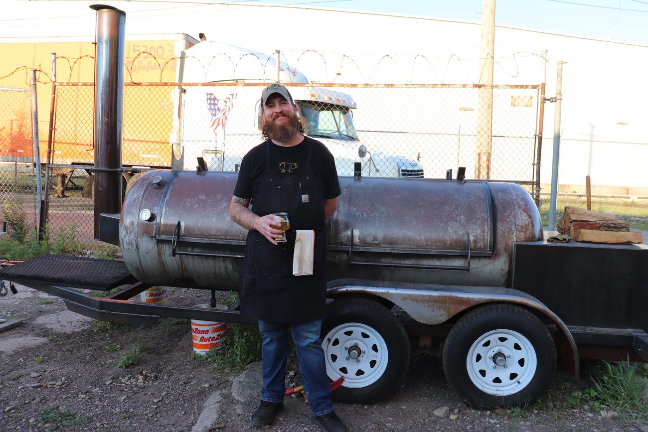 man with large red beard wearing a baseball cap jeans black apron and holding a beer glass leans on a smoker in a dirt patch in front of a chain link fence