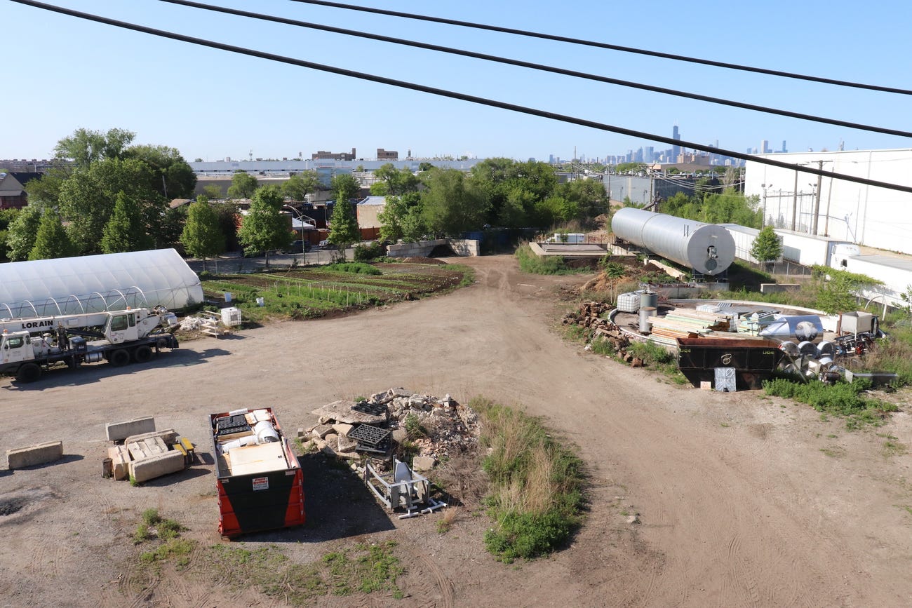 view overlooking a large dirt plot with a greenhouse, a long giant metal cylinder called an anaerobic digester, and rows of crops with the chicago skyline in the distance