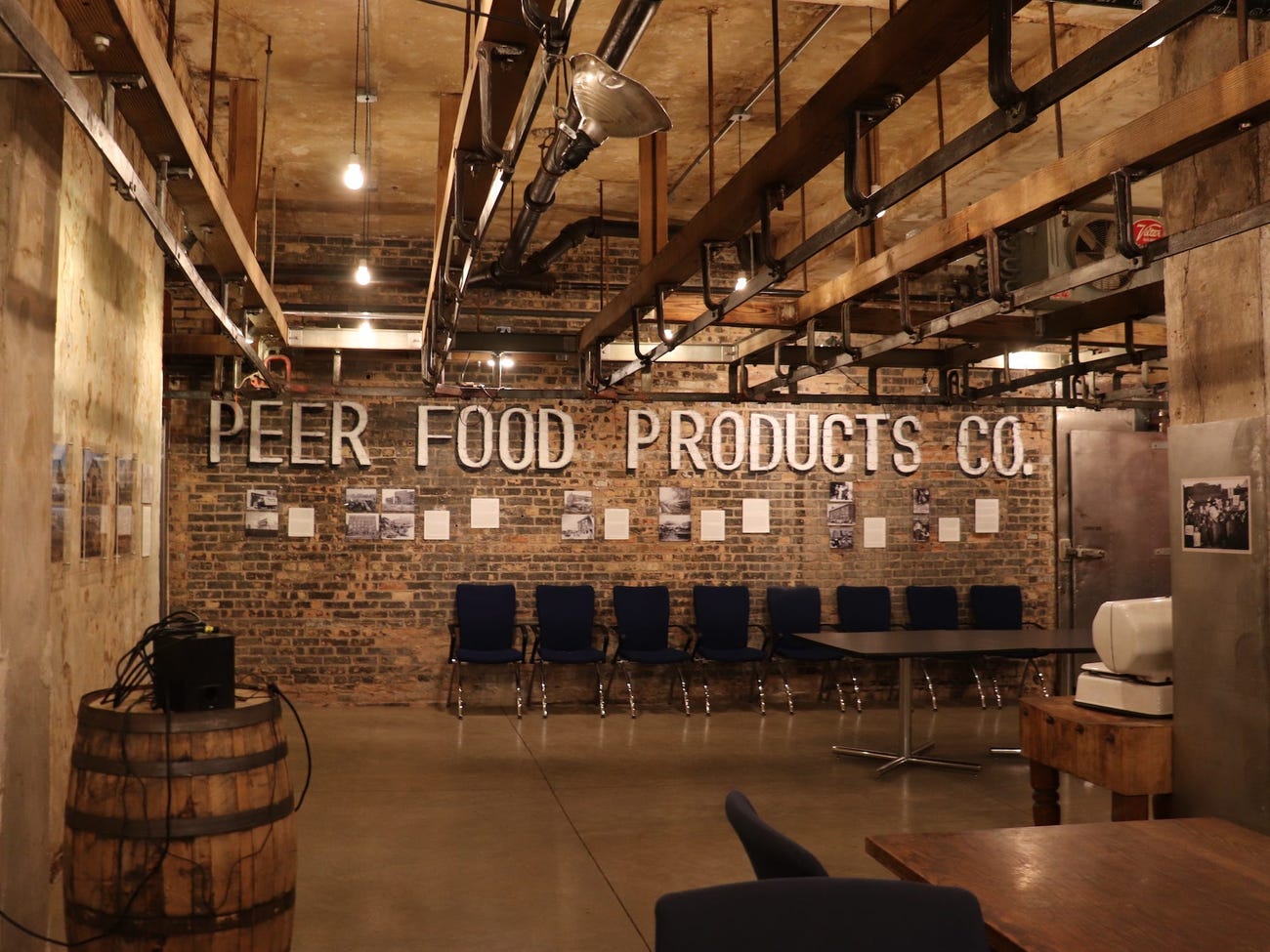 large room with "peer food products co" in white letters on a brick wall with chairs lined up below and old meat railings on the ceiling